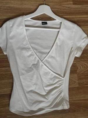 Vit omlottopp med kort ärm från Gina tricot  - Säljer en stilren vit omlottopp med korta ärmar och v-ringning. Toppen har lätt rynkning i sidan och är gjord i mjukt bomullsmaterial. Perfekt till jeans eller kjol för en clean look. Säljer då sen tyvärr inte används längre, pris går att diskutera💕💕