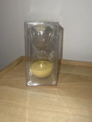 Timglas med guldigt sand - Säljer ett stilrent timglas med guldigt och glittrigt sand. Timglaset är gjort av genomskinligt glas och har en klassisk form. Perfekt som dekoration eller för att hålla koll på tiden vid plugg eller spelkvällar. Har aldrig andvänt den