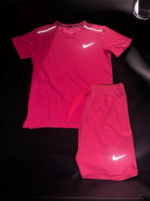 Nike Miler - Snyggt rosa träningsset från Nike med t-shirt och shorts. T-shirten har reflekterande detaljer på axlarna och Nike-logga på bröstet. Shortsen har resår i midjan med dragsko och en reflekterande Nike-logga på benet. Perfekt för gymmet eller löprundan.