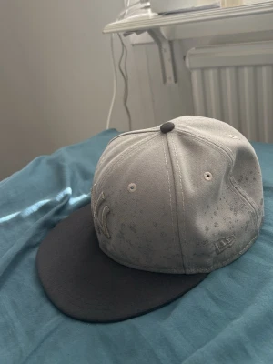 Grå och svart New Era keps - Säljer en stilren keps från New Era i grått med svart skärm. Klassisk modell med broderad logga på sidan och ventilerande hål upptill. Perfekt för dig som vill ha en enkel men snygg accessoar till vardagslooken.