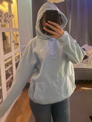 Ljusblå hoodie med glittrig logga - Ljusblå hoodie med huva och stor magficka. Framtill finns en glittrig logga i silver som ger en snygg touch. Hoodien har ribbade muddar vid ärmslut och nederkant. Perfekt för en chill och avslappnad stil.