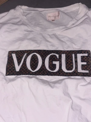 Vit t-shirt med VOGUE-tryck - Säljer en vit t-shirt med ett stort VOGUE-tryck framtill i svart och brunt mönster. T-shirten har rund hals och korta ärmar, perfekt för en trendig och avslappnad stil. Materialet känns mjukt och skönt mot huden.