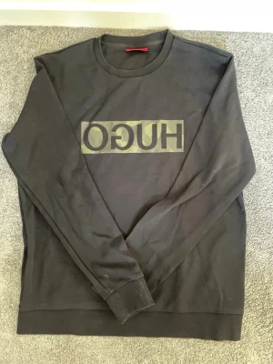 Svart sweatshirt från Hugo Boss - Cool svart sweatshirt från Hugo Boss med stort HUGO-tryck i spegelvänd stil på bröstet. Tröjan har rund halsringning och långa ärmar. Perfekt för en avslappnad och trendig look.