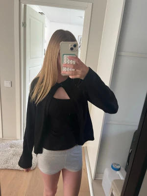 Svart cardigan med cut-out detalj - Snygg svart cardigan med cut-out detalj framtill. Modellen är rak och har långa ärmar. Perfekt att slänga över ett linne eller topp för en stilren look. Enkel att matcha med olika outfits och passar året runt.