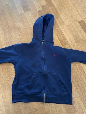 Mörkblå Ralph lauren hoodie med dragkedja - Mörkblå hoodie med dragkedja framtill och kängurufickor. Klassisk design med huva och liten röd logga broderad på bröstet. Tillverkad i mjukt sweatshirtmaterial, perfekt för en avslappnad stil. Stlk 10-12 M