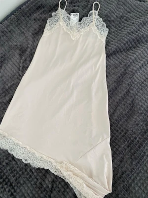 Beige nattlinne med spetsdetaljer - Säljer ett elegant beige nattlinne med tunna axelband och vacker spets vid halsringning och nederkant. Tillverkat i mjukt material med 97% polyester och 3% elastan för en skön känsla. Perfekt för mysiga kvällar hemma.