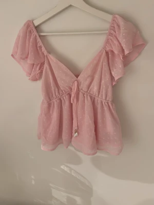 Rosa blus med volang från Amisu - Supersöt rosa blus från Amisu i storlek XS. Blusen har korta volangärmar, knyt framtill och ett diskret prickigt mönster i tyget. Tillverkad i ett lätt och luftigt syntetmaterial, perfekt för varma dagar. Söt och feminin look med v-ringning och flowy passform.