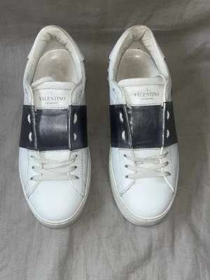 Valentino Garavani vita sneakers - Snygga vita sneakers från Valentino Garavani med bred metallicgrå rem över snörningen. Skorna har klassisk låg profil, vita snören och detaljer i läder. Perfekta för dig som vill ha en clean men ändå lyxig look.