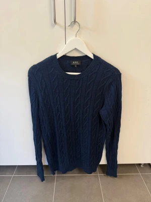 Mörkblå kabelstickad tröja A.P.C. - Snygg mörkblå kabelstickad tröja från A.P.C. med rund halsringning och långa ärmar. Tröjan har ett klassiskt flätmönster och ribbade muddar vid ärmslut och nederkant. Perfekt för dig som gillar stilrena och tidlösa plagg. NYPRIS 3500kr