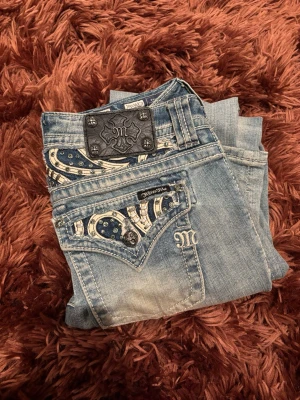 Miss Me ljusblå jeans med paisleydetaljer - Säljer ett par ljusblå jeans från Miss Me med unika paisleymönstrade fickor, dekorerade med nitar och broderier. Jeansen har normal passform med bootcut, låg midja och klassisk femficksdesign. Snygga detaljer på bakfickorna och coolt läderpatch med logga bak❤️❤️Inga defekter vad jag vet, säljer bara pga att dom inte passar på mig💕