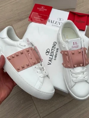 Valentino Garavani vita sneakers - Snygga vita sneakers från Valentino Garavani med ljusrosa band och nitar över vristen. Skorna har en chunky sula med mönstrad undersida och är tillverkade i slätt läder. Perfekta för dig som vill ha en exklusiv och trendig look.