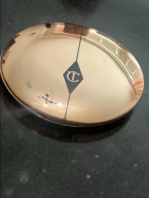 Charlotte Tilbury puderfoundation - Snygg puderfoundation från Charlotte Tilbury i en elegant, roséguldig oval dosa med spegel. Produkten har en ljus beige nyans och ger en silkeslen finish. Perfekt för att jämna ut hudtonen och matta ner glans. Dosan är kompakt och smidig att ta med.