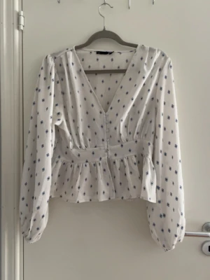 Vit blus med blått blommönster - Söt vit blus med små blå blommor, djup V-ringning och markerad midja. Blusen har långa ärmar med lätt puff och volang nedtill för en romantisk look. Perfekt att styla med jeans eller kjol för en trendig outfit.