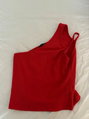 Röd oneshoulder ribbad topp - Snygg röd oneshoulder topp med ribbad struktur. Perfekt för dig som vill sticka ut med en trendig och ungdomlig stil. Toppen är ärmlös och har en smal axelrem på ena sidan.