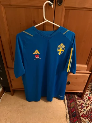 Blå Sverige fotbollströja - Säljer en blå fotbollströja med Sveriges landslagsemblem på bröstet och gula detaljer på ärmarna. Tröjan har V-ringning och korta ärmar, samt tryck från Svenska Spel och Adidas på framsidan. Perfekt för match eller träning.