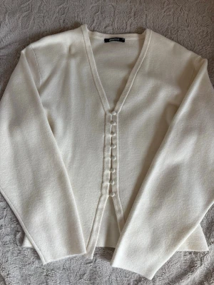 Vit kofta från Gina Tricot - Stilren vit kofta från Gina Tricot med v-ringning och dekorativa knappar framtill. Koftan har långa ärmar och är gjord i ett mjukt, stickat material som känns skönt mot huden. Perfekt att slänga över en topp för en enkel men snygg look.