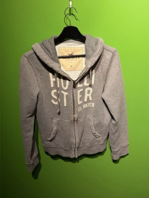 Grå zip hoodie från Hollister - Snygg grå hoodie från Hollister med dragkedja framtill och stora fickor. Tryckt logga i vitt på bröstet och justerbar huva med snören. Mjuk insida och ribbade muddar vid ärmar och nederkant. Perfekt för en avslappnad och cool stil.