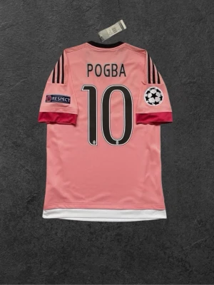 Juventus Pogba matchtröja rosa - Juventus matchtröja i rosa med Pogba och nummer 10 på ryggen. Tröjan har svarta Adidas-ränder på axlarna, Juventus klubbmärke och Jeep-logga på bröstet samt Champions League-märke på ärmen. Tillverkad i lätt och ventilerande material.