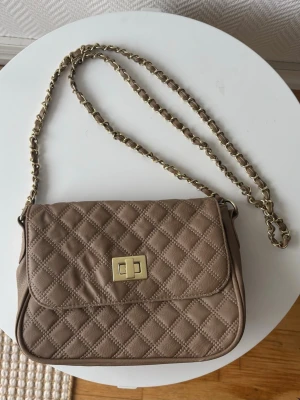 Beige quiltad axelväska med kedja - Taupe - Snygg beige axelväska med quiltat mönster och guldfärgad kedjerem. Väskan har ett vridlås i guld på framsidan och en mjuk, rektangulär form. Perfekt för dig som gillar trendiga accessoarer och vill ha plats för det viktigaste.