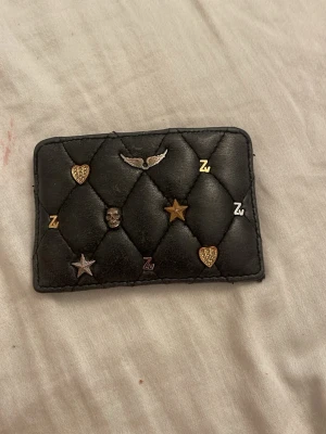 Zadig & Voltaire plånbok quiltad  - Säljer en svart quiltad plånbok i läderimitation med coola metalldetaljer som stjärnor, hjärtan, dödskalle och bokstäver. Plånboken har ett kompakt format och plats för kort. Perfekt accessoar för dig som gillar edgy detaljer och unik stil. Den är från Zadig köpt för ett år sen, i princip i nytt skick 