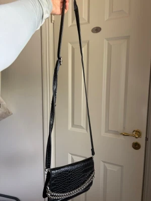 Svart axelväska med kedjedetalj - Snygg svart axelväska i croco-mönstrat konstläder med justerbar axelrem och en silverfärgad kedja framtill. Väskan har en stilren och modern look som passar perfekt till olika outfits. Perfekt storlek för det viktigaste och enkel att bära över axeln.