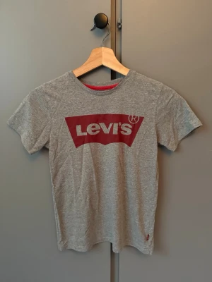 Grå Levi's t-shirt med röd logga - Snygg grå t-shirt från Levi's med klassisk röd logga framtill. T-shirten har rund halsringning och korta ärmar. Tillverkad i mjuk bomull som känns skön mot huden. Perfekt för en avslappnad och stilren look.