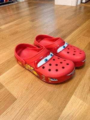 Röda Crocs Cars tofflor - Säljer ett par röda Crocs tofflor med motiv av Blixten McQueen från Disney Cars. Tofflorna har perforerad ovandel, justerbar hälrem och detaljer som ögon och nummer 95 på sidorna. Tillverkade i lätt och bekvämt syntetmaterial. Lightning mcqueen 