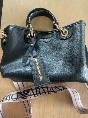 Svart handväska Emporio Armani - Stilren svart handväska från Emporio Armani i präglat läder med guldfärgade metalldetaljer. Väskan har dubbla handtag, avtagbar axelrem med logga och en dekorativ tag med Emporio Armani-tryck. Perfekt för dig som gillar klassisk och lyxig design.
