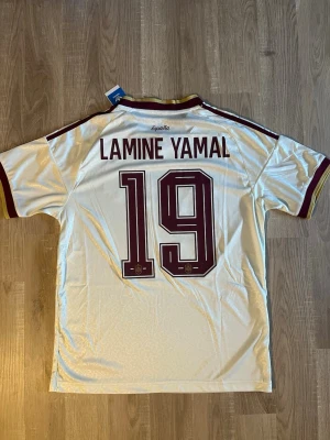 Vit Spanien fotbollströja Lamine Yamal  - Säljer en vit Spanien fotbollströja med vinröda detaljer och nummer 19. Tröjan har LAMINE YAMAL tryckt på ryggen, klubbmärke på bröstet och klassiska ränder på axlarna. Tillverkad i ett lätt och ventilerande material, perfekt för träning eller match.