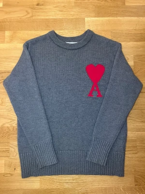 Ami Paris S - Mysig grå stickad tröja med rund hals och ribbade muddar. På bröstet finns ett stort rött hjärta och bokstaven A i samma färg, vilket ger en unik och trendig look. Perfekt för dig som gillar stilrena plagg med en twist.