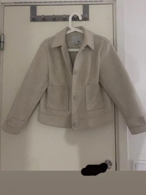 Beige ullkappa från 157 - Stilren beige ullkappa från 157 med klassisk krage och stora framfickor. Kappan har knäppning framtill och på ärmsluten samt en rak, något boxig passform. Perfekt för dig som gillar minimalistisk och tidlös stil. Ganska stor storleken på affären nog folk med större storlekar