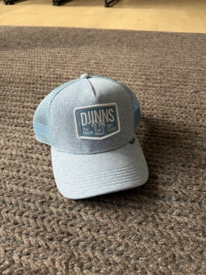 Kepsar - Snygg ljusblå truckerkeps från DUNNS med mesh på sidorna och broderad patch framtill där det står 'DUNNS Do Nothing Club'. Skön passform och justerbar baktill. Perfekt för en avslappnad streetwear-look.