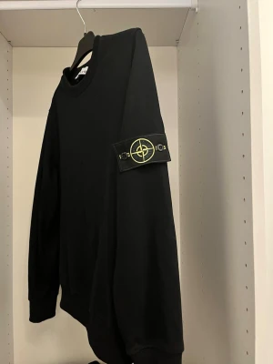 Stone Island tröja - Säljer en svart sweatshirt från Stone Island med den ikoniska kompasspatchen på vänster ärm. Tröjan har rund halsringning, ribbade muddar och är tillverkad i mjukt material. Perfekt för dig som gillar streetwear och stilrena plagg.