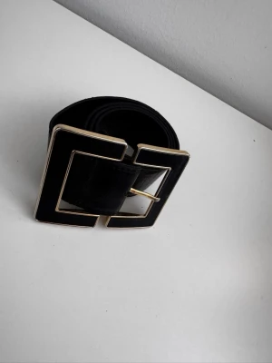 Svart bälte med stor fyrkantig spänne - Snyggt svart bälte med ett stort, fyrkantigt spänne i guldtonad metall. Bältet har en clean och modern look som passar till många olika outfits. Tillverkat i skinn för en exklusiv känsla och extra hållbarhet.