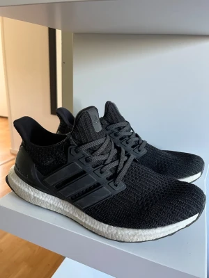 Svarta Adidas Ultra Boost sneakers - Säljer ett par svarta Adidas Ultra Boost sneakers med stickad ovandel och vita Boost-sulor. Skorna har svarta plastdetaljer och snörning samt diskret Ultra Boost-logga på hälen. Perfekta för dig som gillar stilrena och bekväma sneakers.