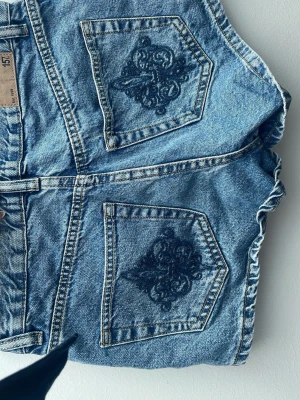 Blå denimshorts med broderade fickor - Säljer ett par blå låg midjade denimshorts med snygga broderade detaljer på bakfickorna. Shortsen har normal passform, klassisk femficksdesign och bälteshällor. Perfekta för en avslappnad och trendig look i sommar.