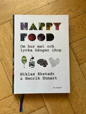 Happy Food av Ekstedt & Ennart - En inspirerande bok med hårda pärmar som utforskar sambandet mellan mat och lycka. Omslaget är vitt med färgglada bokstäver och illustrationer av grönsaker, hjärna och hjärta. Perfekt för dig som vill förstå hur kosten påverkar välmående.