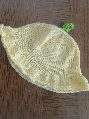 Gul stickad mössa med blad - Söt handstickad mössa i ljusgul färg med ett litet grönt blad på toppen. Mössan har en mjuk och lite lös passform och är tillverkad i tunt garn, vilket ger en lätt och bekväm känsla. Perfekt för dig som vill ha en unik accessoar med lekfull touch.