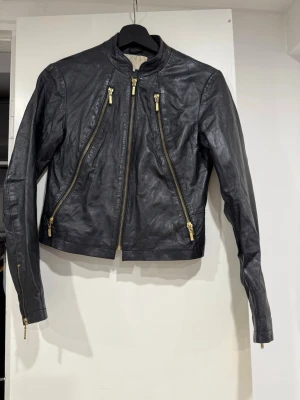 Svart skinnjacka med guldiga dragkedjor - Snygg svart skinnjacka i bikerstil med fyra guldiga dragkedjor framtill och ståkrage. Jackan har en kortare passform och långa ärmar med dragkedjor vid ärmsluten. Perfekt för dig som vill ha en edgy och trendig look.