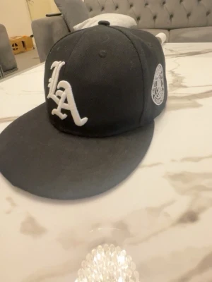 Svart LA snapback keps - Säljer en svart snapback keps med stor vit LA-logga framtill och ett broderat märke på sidan. Kepsen har platt skärm och justerbar baksida med plastspänne. Perfekt för dig som gillar streetwear och vill ha en stilren accessoar.