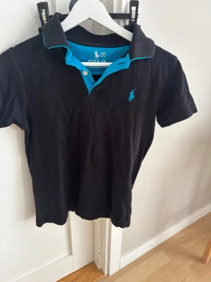 Svart pikétröja från Polo Ralph Lauren - Klassisk svart pikétröja från Polo Ralph Lauren med turkosa detaljer och logga på bröstet. Tröjan har korta ärmar, krage och knappslå framtill. Perfekt för en avslappnad men ändå stilren look. Har ett litet hål som man inte märker irl men ja tog bild på de