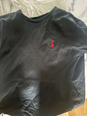 Svart t-shirt från Polo Ralph Lauren, - Klassisk svart t-shirt från Polo Ralph Lauren med röd broderad logga på bröstet. Modellen är custom slim fit och har korta ärmar samt rund halsringning. Tillverkad i mjuk bomull som känns skön mot huden. Den är M men är kort och mer som S