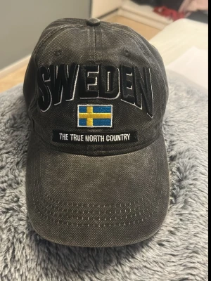 Grå keps med SWEDEN-brodyr - Säljer en grå keps med stor brodyrtext 'SWEDEN' framtill, svensk flagga och texten 'THE TRUE NORTH COUNTRY' under. Kepsen har böjd skärm och justerbar baksida. Materialet ser ut att vara bomullstyg med tvättad look. Perfekt för dig som vill visa svensk stolthet.