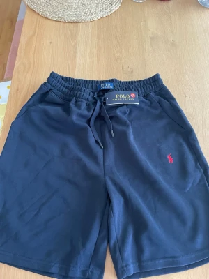 Polo Ralph Lauren shorts - Svarta shorts från Polo Ralph Lauren med elastisk midja och dragsko. Klassisk röd logga broderad på vänster ben. Tillverkade i mjukt bomullsmaterial och har två sidofickor. Perfekta för en avslappnad och stilren look. Riktigt sköna. Storlek M. Kom privat vid minsta funderingar!