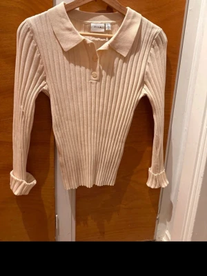 Beige ribbad tröja med krage - Snygg beige ribbstickad tröja med klassisk krage och knäppning framtill. Tröjan har långa ärmar och är figurnära i modellen, perfekt för en stilren look. Materialet är mjukt och stretchigt för extra komfort.