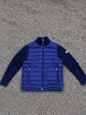 Cardigan - Snygg blå pufferjacka med quiltad front och stickade ärmar. Jackan har hög krage, två dragkedjefickor och en M0ncIer-logga på ärmen. Perfekt för kalla dagar och ger en sportig men stilren look.