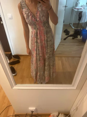 Lång bohemisk maxiklänning med volang xs-s - Säljer en lång maxiklänning med bohemiskt mönster i rosa, beige och grå toner. Klänningen har v-ringning med volangkant, smock i midjan och axelband med volangdetaljer. Aldrig använd