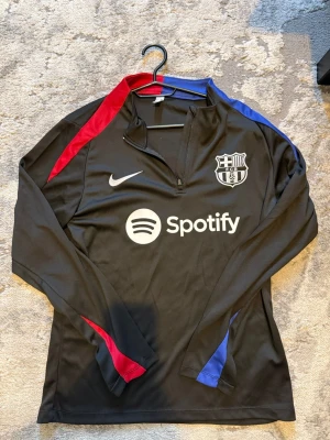 Svart FC Barcelona träningströja Nike - Snygg svart långärmad träningströja från Nike med FC Barcelona-logga och Spotify-tryck på bröstet. Tröjan har dragkedja vid halsen samt röda och blå detaljer längs ärmarna. Perfekt för fotbollsträning eller som supporterplagg.