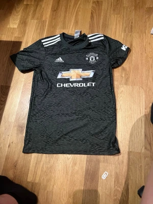 Manchester United bortatröja Adidas - Snygg svart Manchester United bortatröja från Adidas med vita detaljer och tre ränder på axlarna. Tröjan har Manchester Uniteds klubbmärke och Chevrolet-logga på bröstet. Tillverkad i lätt och ventilerande material, perfekt för fotboll eller träning.