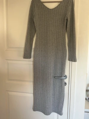 Grå ribbad lång stickad klänning - Långärmad grå stickad klänning med ribbad struktur och v-ringning. Klänningen är lång och har en kroppsnära passform som framhäver siluetten. Perfekt för dig som gillar en enkel men stilren look.